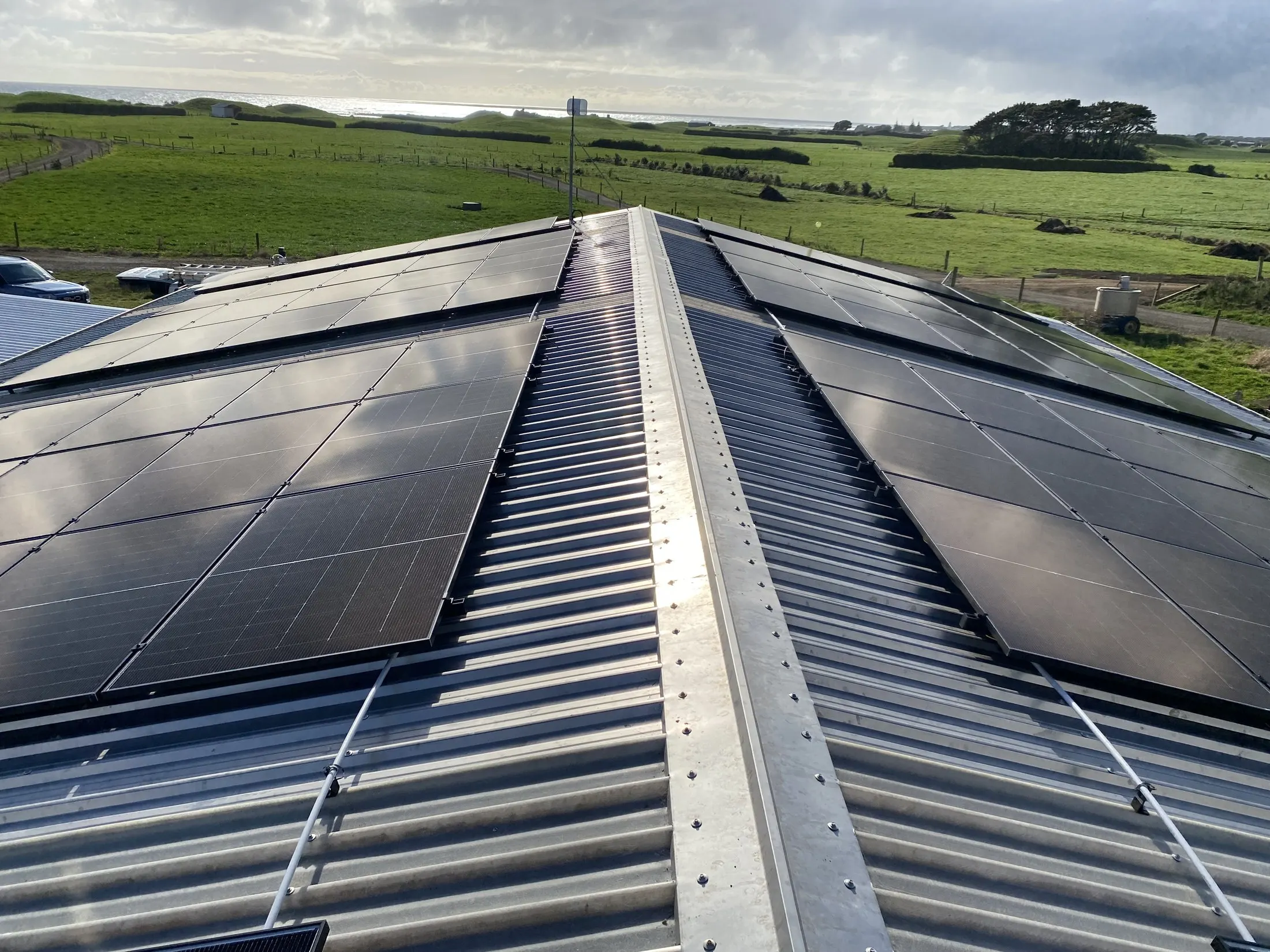 Solar arrays on both sides of a dairy shed roof ridge with coastal farmland and ocean beyond, Pungarehu